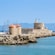 A stone fortress with a lighthouse and 2 windmills on a rocky pier, surrounded by sailboats and calm water